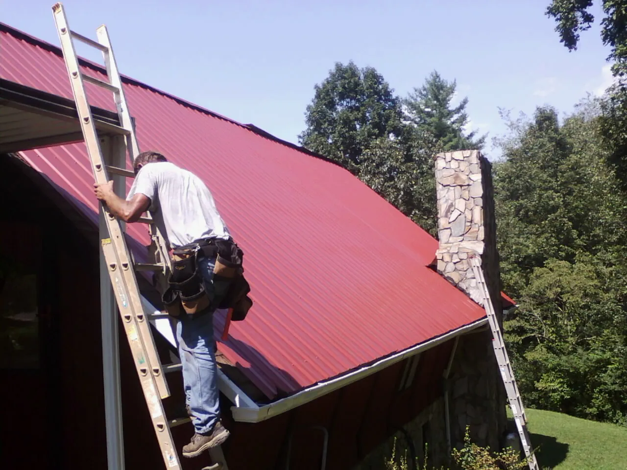 Roofer on a ladder beside a red metal roof and stone chimney — Soffit & Fascia Repair by Roofing Brothers in Alderson